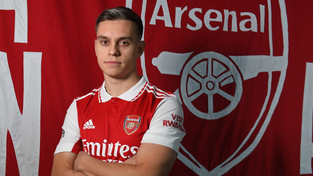 Leandro Trossard posing cross-armed in front of an Arsenal flag.