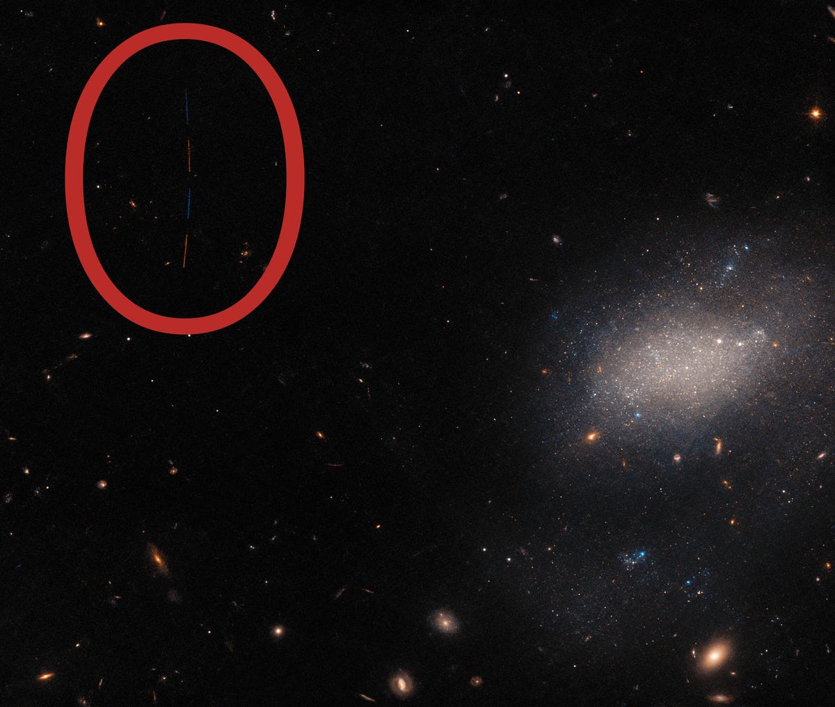 Dark space with a diffuse, hazy galaxy in the middle, distance galaxies in the background and a small trail of a streaking asteroid. Rec circle around the asteroid trail.