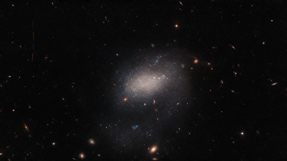 Dark space with a diffuse, hazy galaxy in the middle, distance galaxies in the background and a small trail of a streaking asteroid.