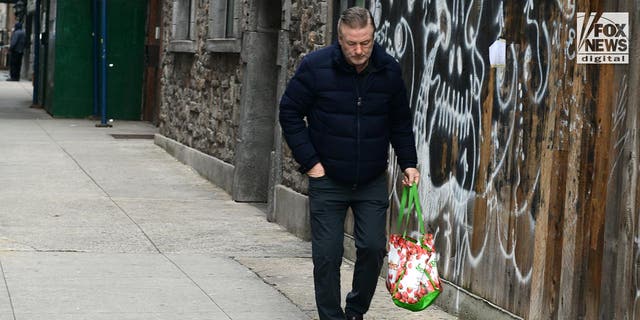 Alec Baldwin dressed in dark clothes has his hand in his pocket as he walks along the sidewalk