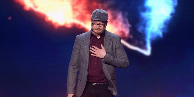 Art Garfunkel Jr. wearing a gray blazer and matching hat with a burgundy shirt performing on stage