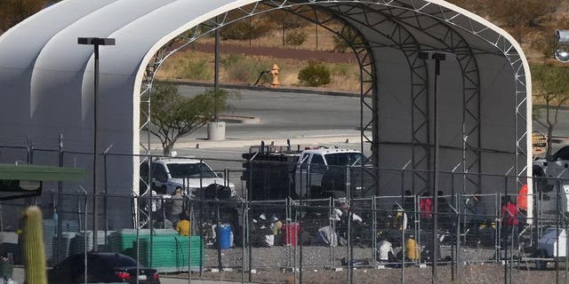 Migrants sit on the ground behind fencing under a fabric arched canopy b