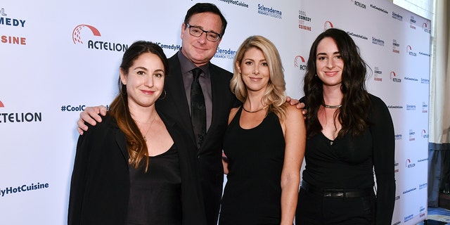 Aubrey Saget in all black stands next to father Bob Saget in a black suit and tie, next to wife Kelly Rizzo in a black dress, next to Lara Saget, also in all black on the red carpet