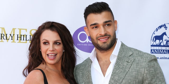 Britney Spears in a black dress looks to her right in a black dress as Sam Asghari looks to his left in a grey suit