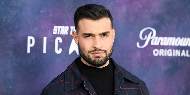 Sam Asghari soft smirks on the carpet in a dark plaid jacket and black shirt