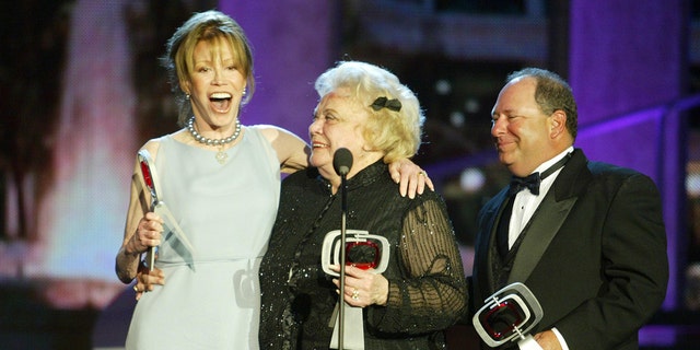 Mary Tyler Moore in a pale green sleeveless dress being embraced by Rose Marie wearing a black sheer dress