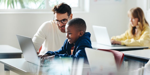 Male teacher helps a young boy with computer-based learning in a classroom setting.