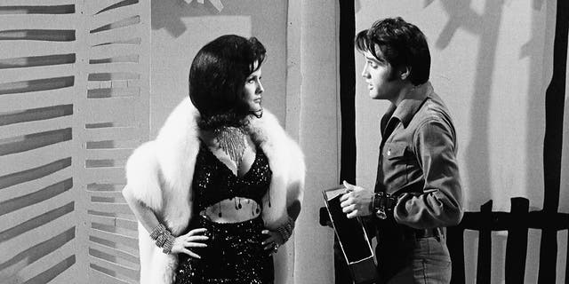 A model wearing a bejeweled bra top and skirt in a white fur coat talking to Elvis Presley in a denim suit