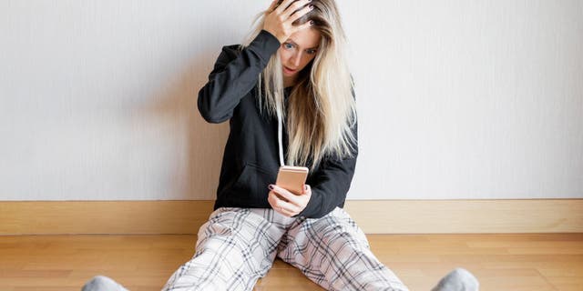 Woman places hand on head in shock with eyes wide and mouth open looking down at her phone