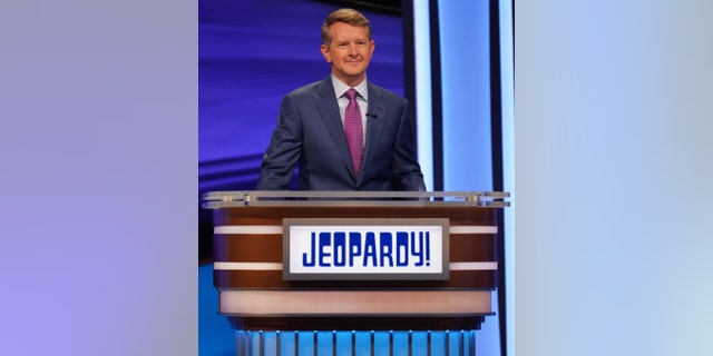 Ken Jennings in a blue suit with a purple tie behind the Jeopardy! podium