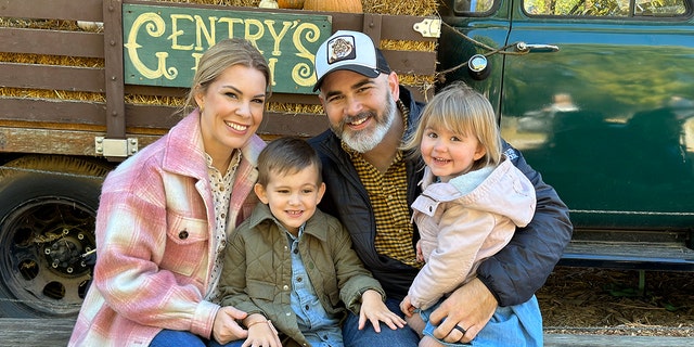 Tasha Layton wearing a pink plaid jacket, a printed blouse and jeans posing with her smiling husband and two children