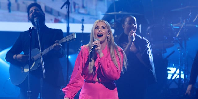 Tasha Layton wearing a hot pink jumpsuit and singing on a mic
