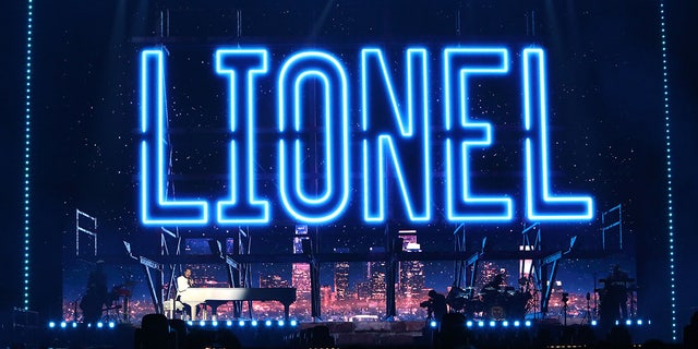 Lionel Richie on stage playing from a white piano with his name outlined in blue lights on the screen behind himawan