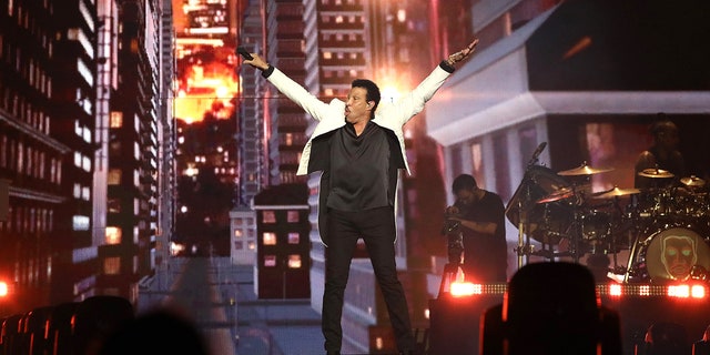 Lionel Richie wears a white jacket and black pants and shirt and puts his arms in the air on his tour with Earth, Wind & Fire