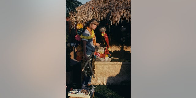 Michelle Pfeiffer wearing leis over a grey sweatshirt, with a gold-coloured grass skirt over a pair of jeans, holding a model of a red parrot while standing beside a thatched hut, on the set of Grease 2