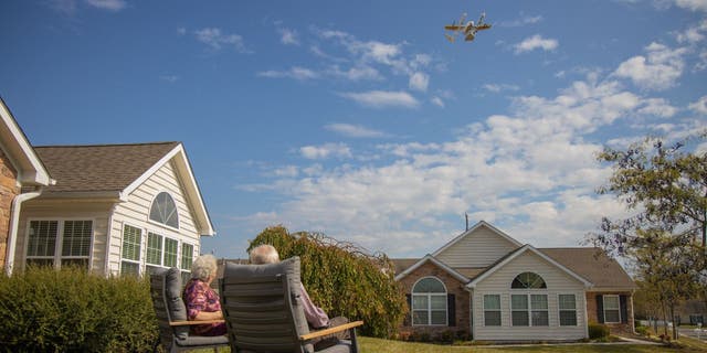 Susie and Paul sitting in chairs outside as a drone in the sky brings them their order