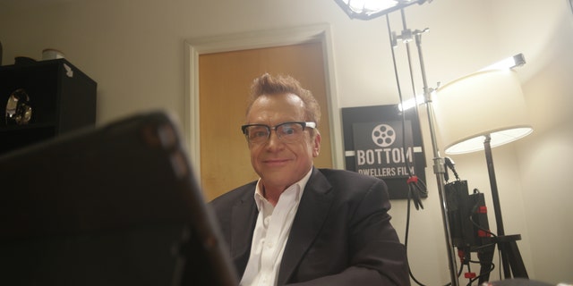 Tom Arnold sitting at a desk in character