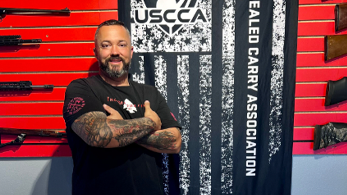 Joe Talachy poses by wall holding guns, USCCA flag