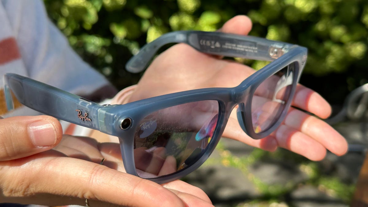 Hands holding a gray pair of sunglasses with cameras in the corners of the frames, made by Meta and Ray-Ban