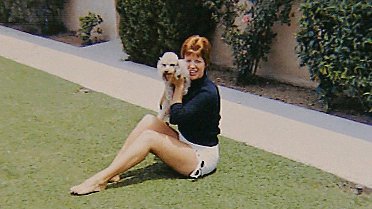 Ruth Marie Terry wearing a blue sweater and white shorts sitting on the grass and holding a white dog.