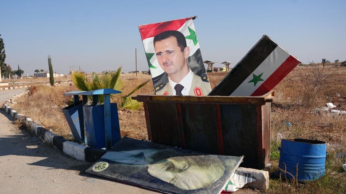 TOPSHOT - A picture taken at the entrance of the Kweyris military airfield in the eastern part of Aleppo province on December 3, 2024 shows a portrait of  Syria's President Bashar al-Assad and a national flag in the garbage dumpster following the take over of the area by rebel groups. A war monitor on December 1 said Ankara-backed groups seized control of the towns of Safireh and Khanasser southeast of Aleppo from government forces, and also took the Kweyris military airport.