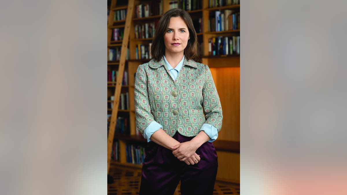 Amanda Knox wearing a green blazer, a light blue shirt and dark pants standing in front of a library.