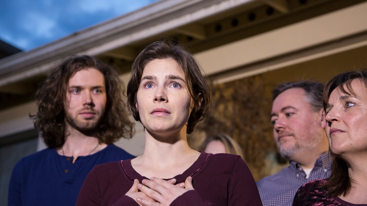 Amanda Knox with her hands to her chest with people behind her.