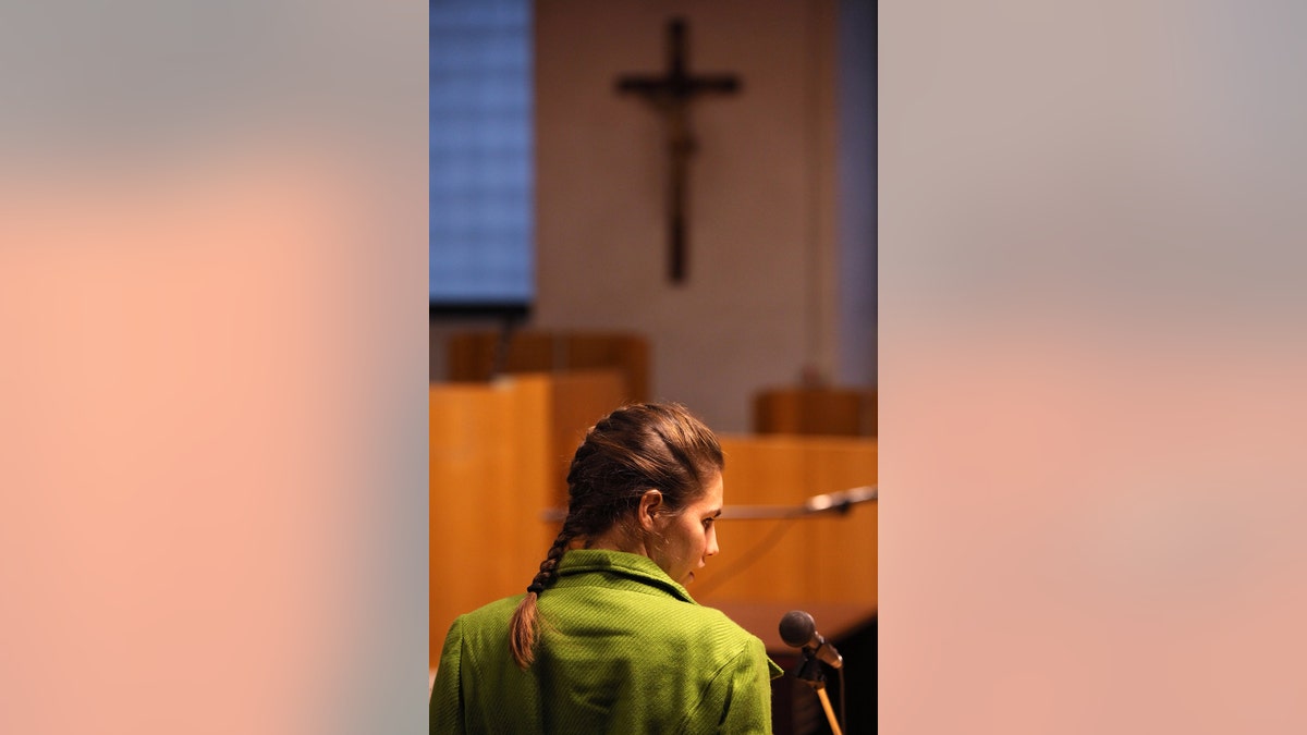 Amanda Knox with her back turned in a green jacket facing a cross.