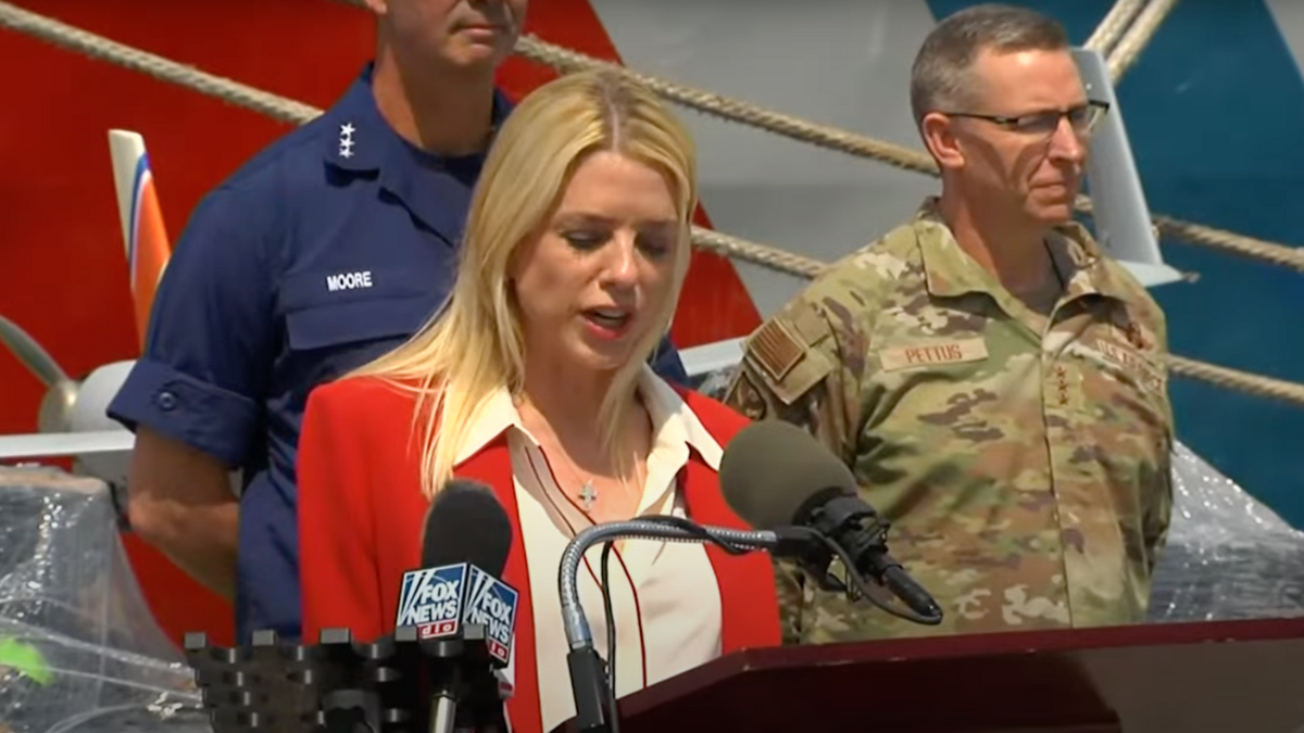 Pam Bondi wears a red jacket while speaking at a press conference in front of a Coast Guard ship.