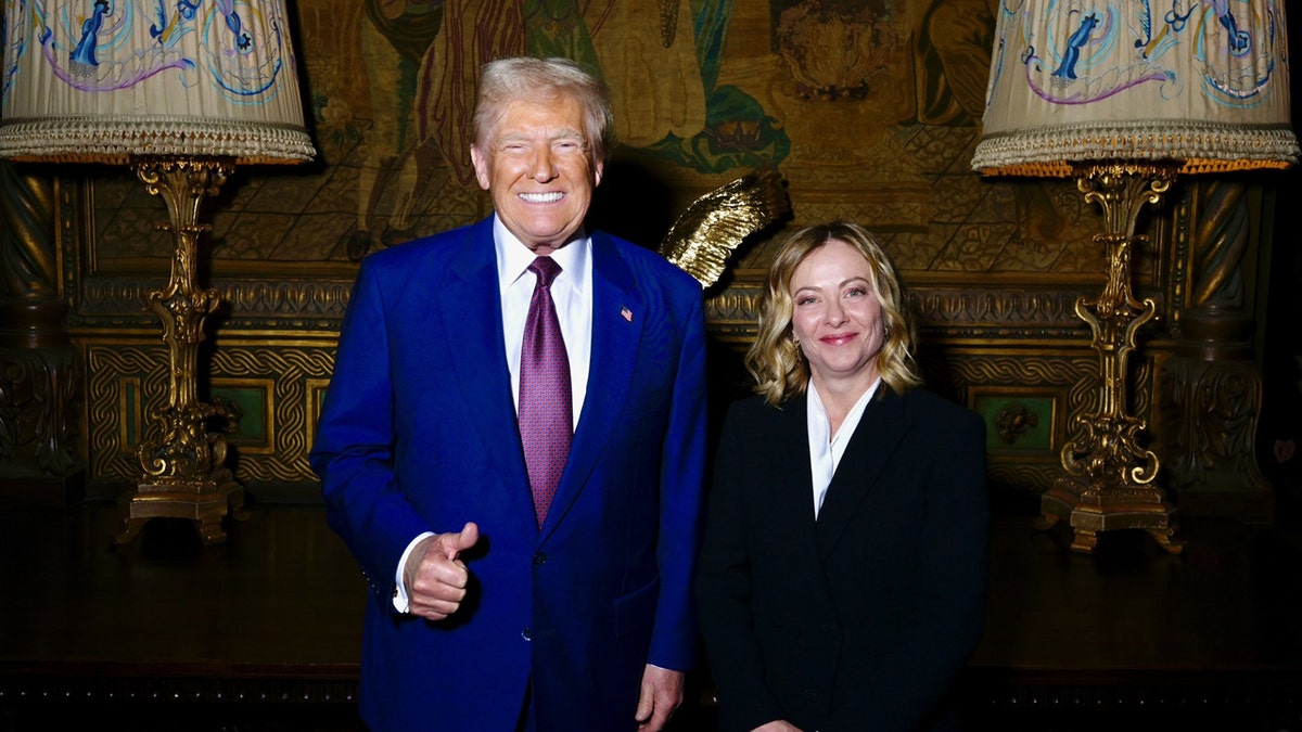 President-elect Donald J. Trump gives a thumbs up in a blue suit as he stands next to Italian Prime Minister Giorgia Meloni at his Mar-a-Lago residence. Both are smiling.
