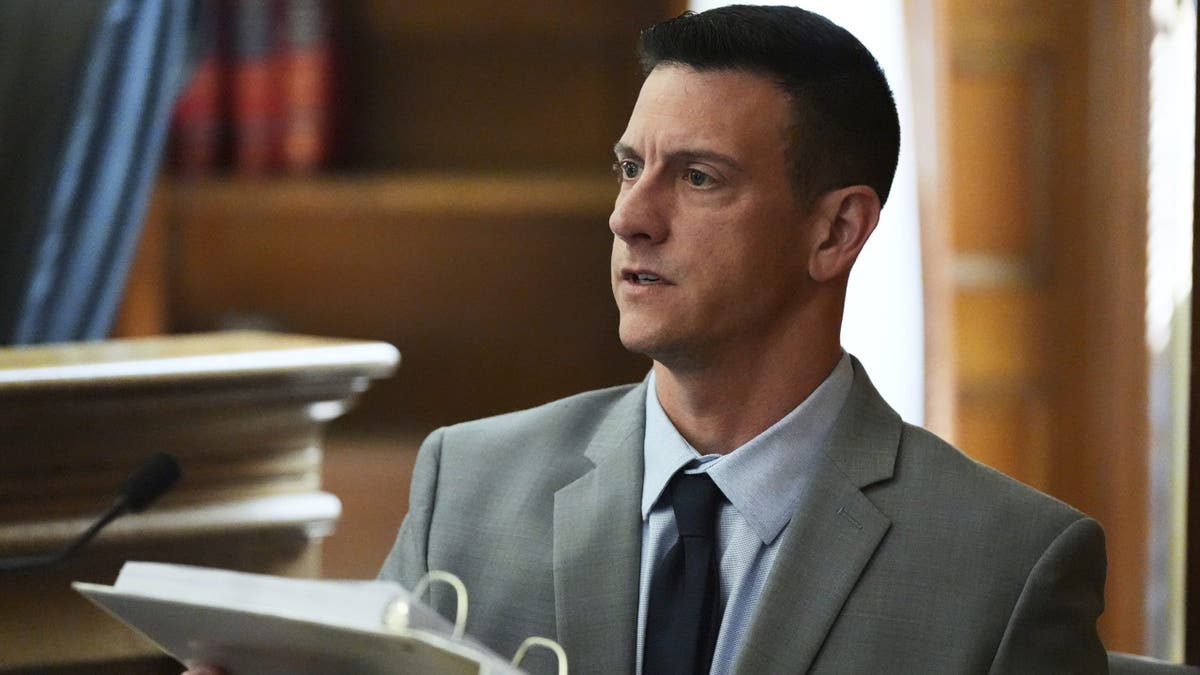 Mass. State Police detective Nicholas Guarino wearing a gray suit and holding a binder that contains text messages between Karen Reade and John O'Keefe while on the witness stand at her retrial on murder charges