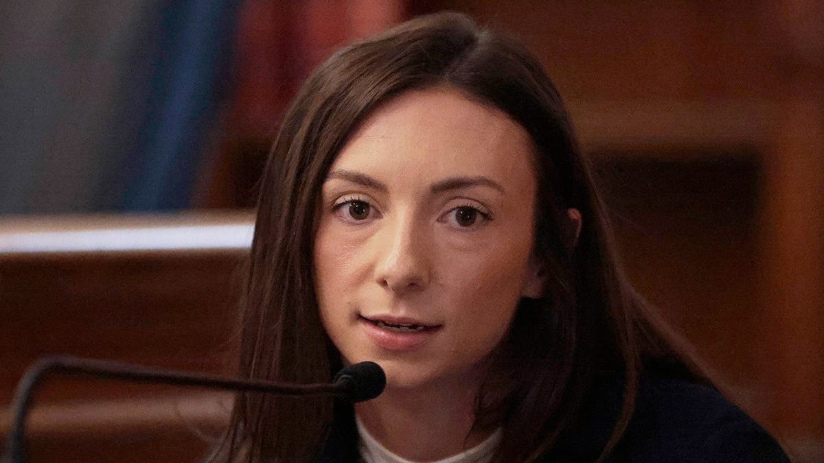 Katie McLaughlin, wearing a dark jacket, white shirt and gold necklace, leans into the microphone from the witness stand