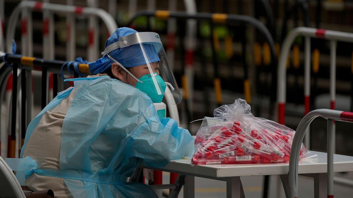 Volunteer wears protective gear sitting down at table with coronavirus testing kits on table in Beijing