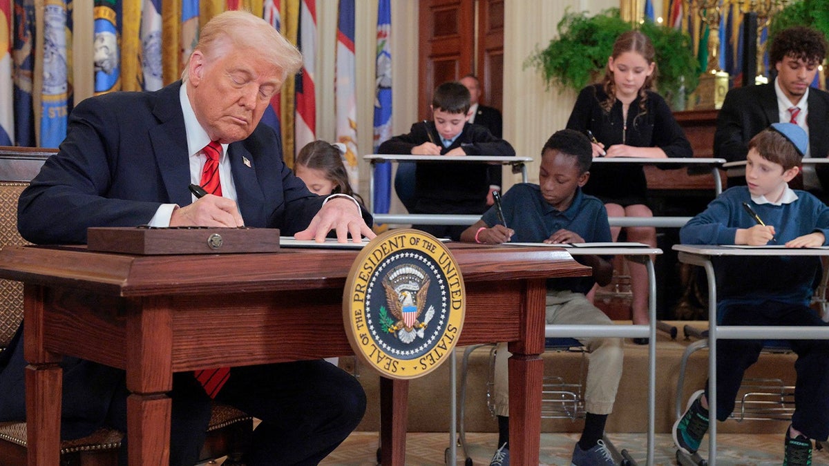 Trump signing document at desk, kids to his left at desks