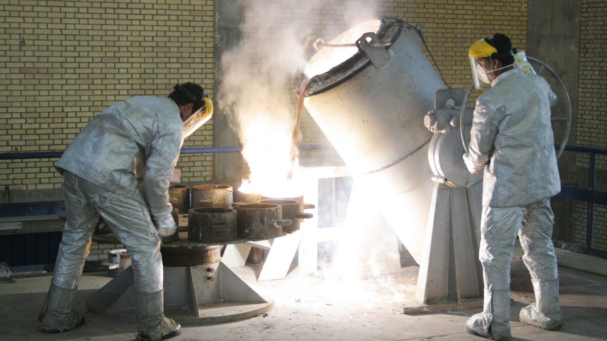 Two technicians in reflective protective suits and face shields operate equipment to pour molten material inside a uranium conversion facility near Isfahan, Iran, on March 30, 2005. Steam and light radiate from the process as they work near a reactor-like container.