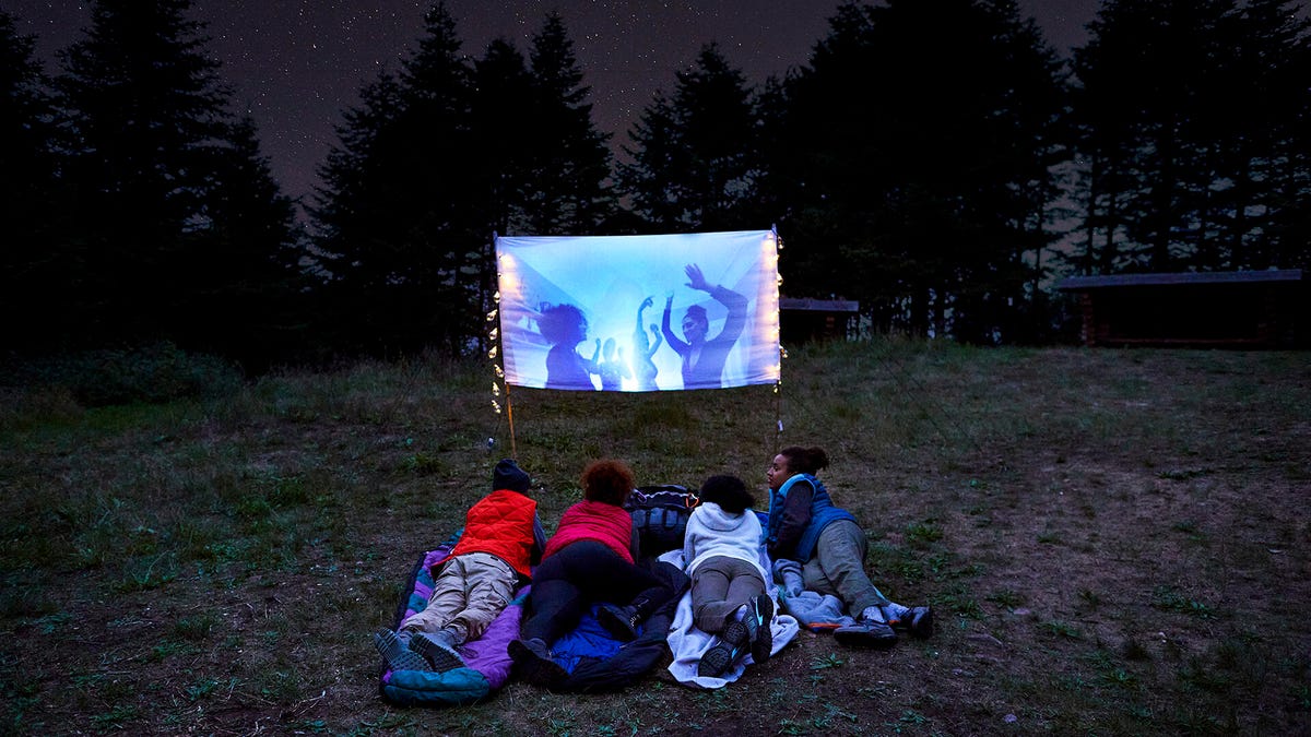Four kids lay in the grass under the stars watching a movie on a small screen.