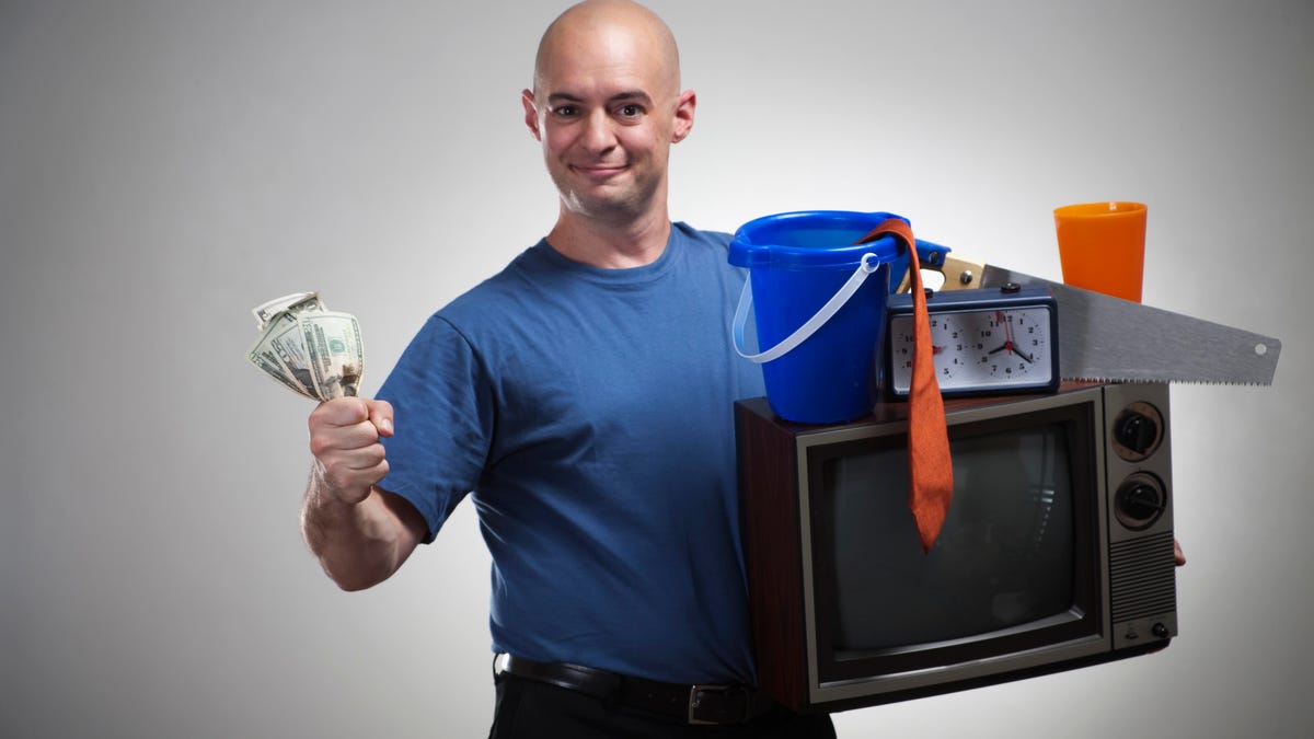A bald man in a blue shirt holds cash in one hand and a TV with some junk on it in the other.