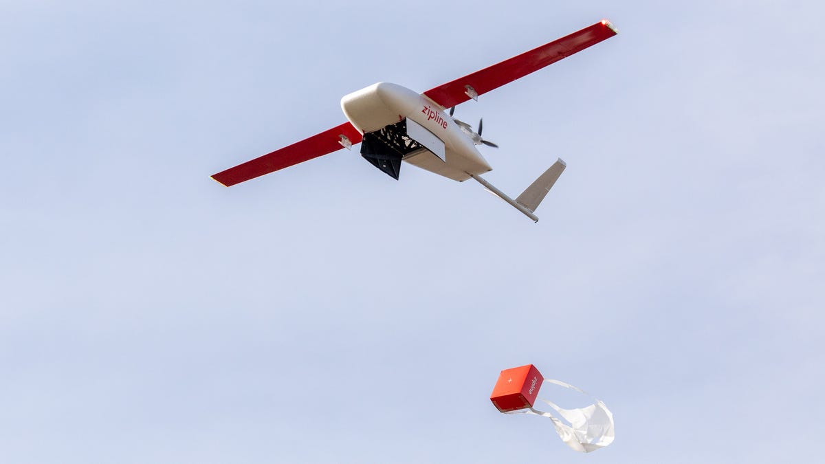 A drone like a small airplane drops a red box, its parachute unfurling, out of a hatch.