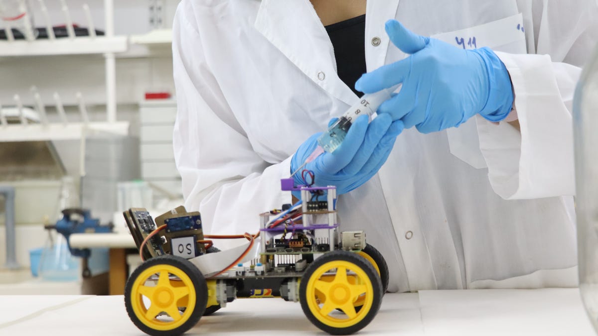 A small tabletop robot is seen with yellow wheels. A scientists is injecting a liquid into one area of the robot.