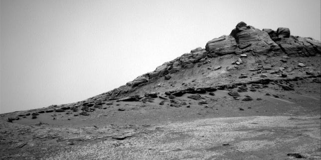 Black and white rocky Mars landscape with blocky hill and a thin layer of darker rocks running partway up it.