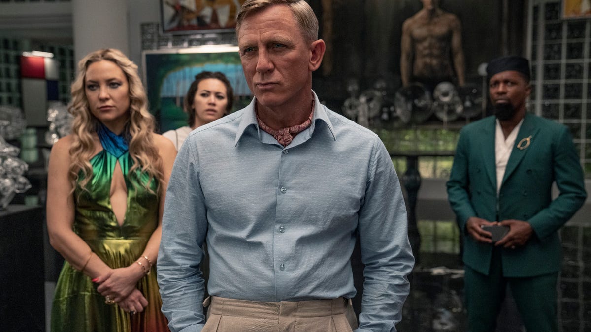Benoit Blanc, played by Daniel Craig, stands with his hands in his pockets and an inscrutable look on his face, with other characters from the film standing in the background behind him.