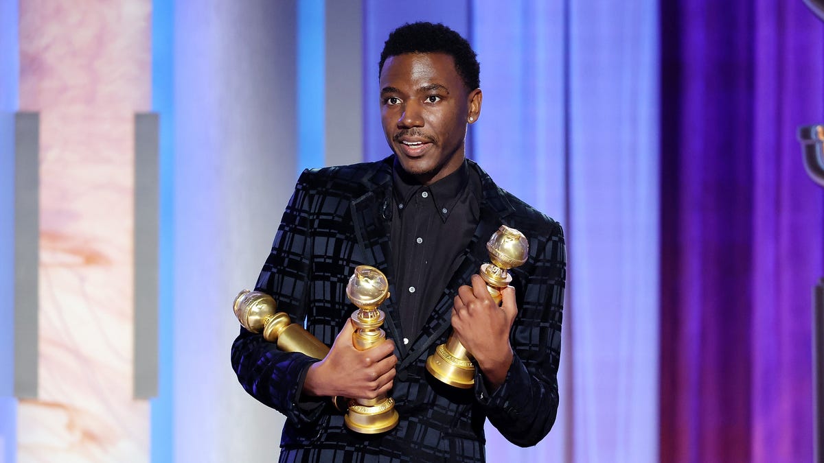 Host Jerrod Carmichael on stage in a black suit holding an armful of Golden Globe Awards.