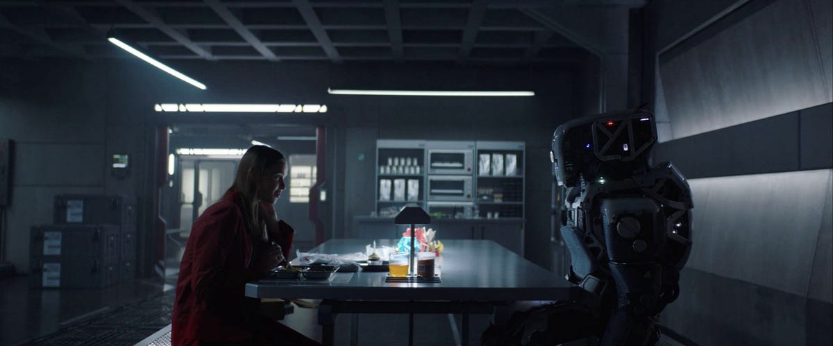 A robot sits on the right, across a table from a girl who is on the left.