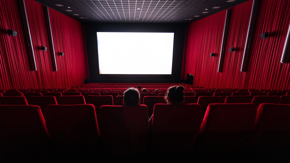Two people sit in a theater auditorium with deep red walls facing a shining white screen