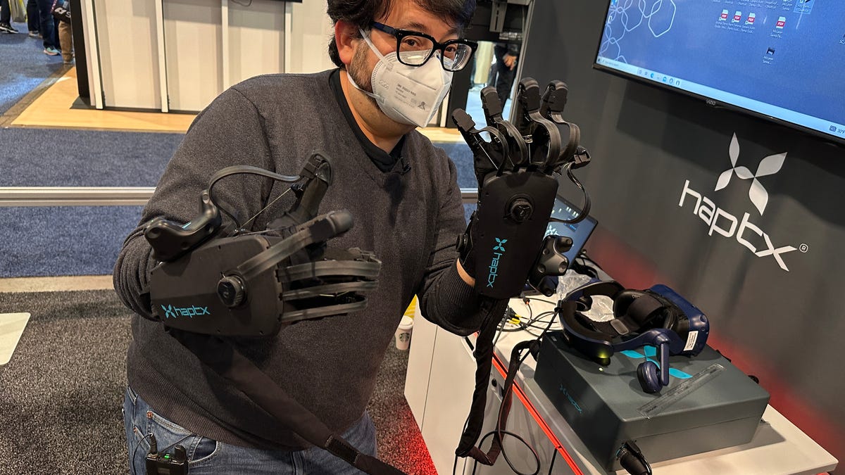 A man wearing large wired haptic gloves in a room next to a computer