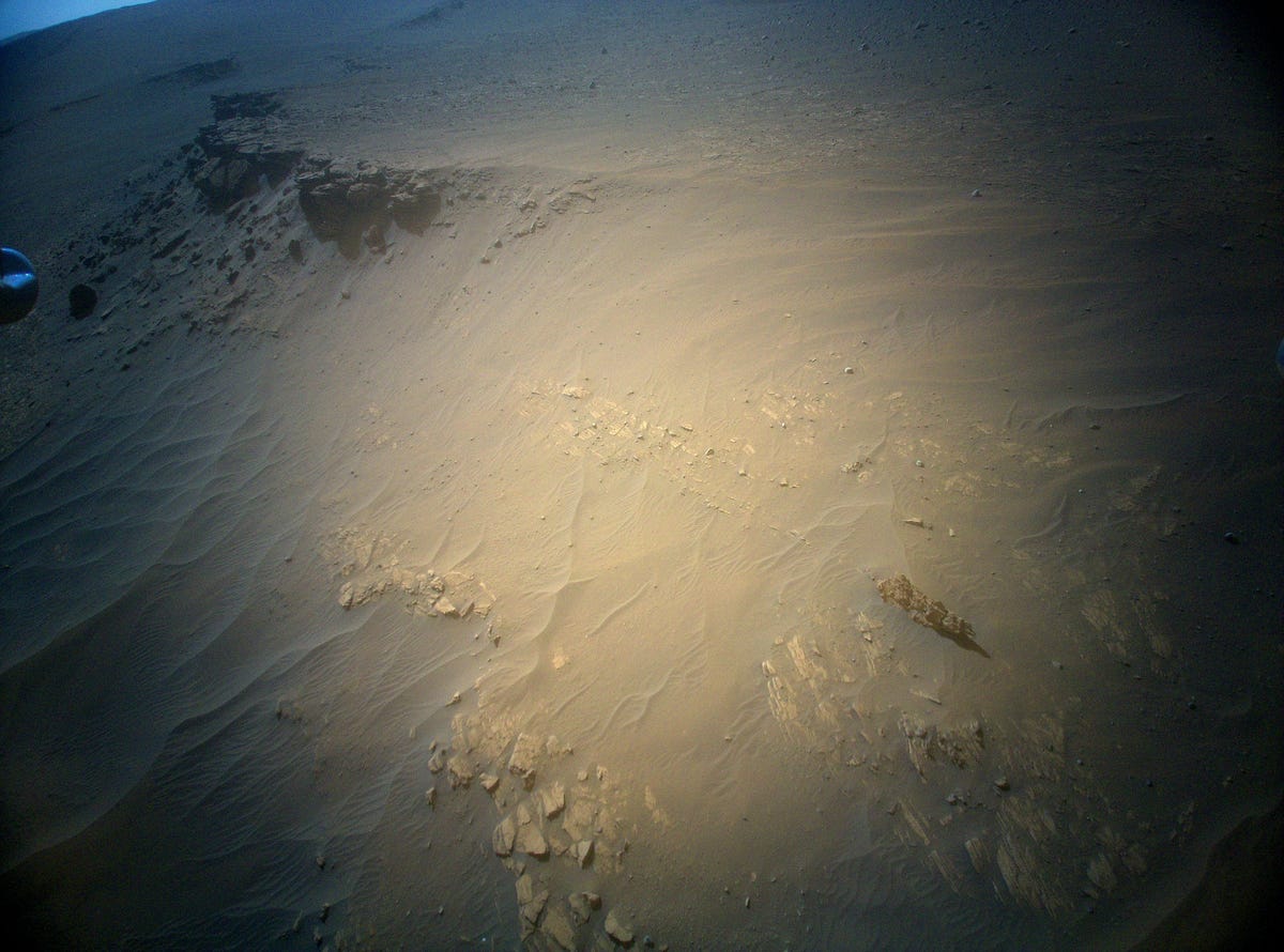 Ingenuity helicopter view of a low, rocky ridge and sandy Martian ground. A tiny sliver of sky is visible in the corner.
