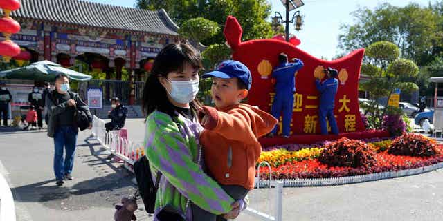 A woman wearing a mask carries a child past workers decorating a display celebrating the upcoming 20th Party Congress in Beijing.
