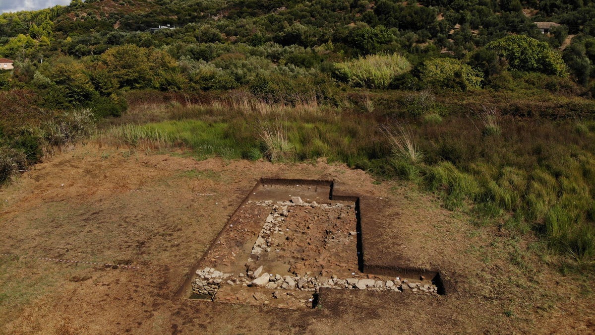 Excavation site on Greek coast shows a green hill in the background with a geometric structure partially uncovered on the ground.