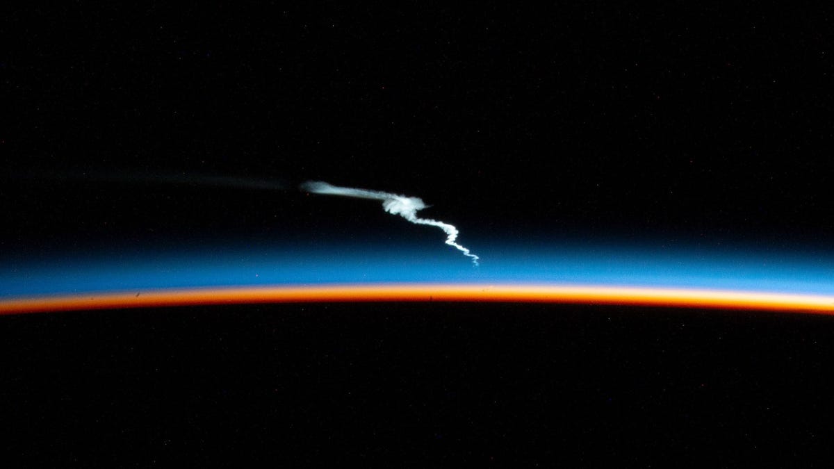 Glowing blue and orange curve of Earth with darkness below and darkness of space above. A swirling white plume from a Falcon Heavy rocket launch slashes across the middle.