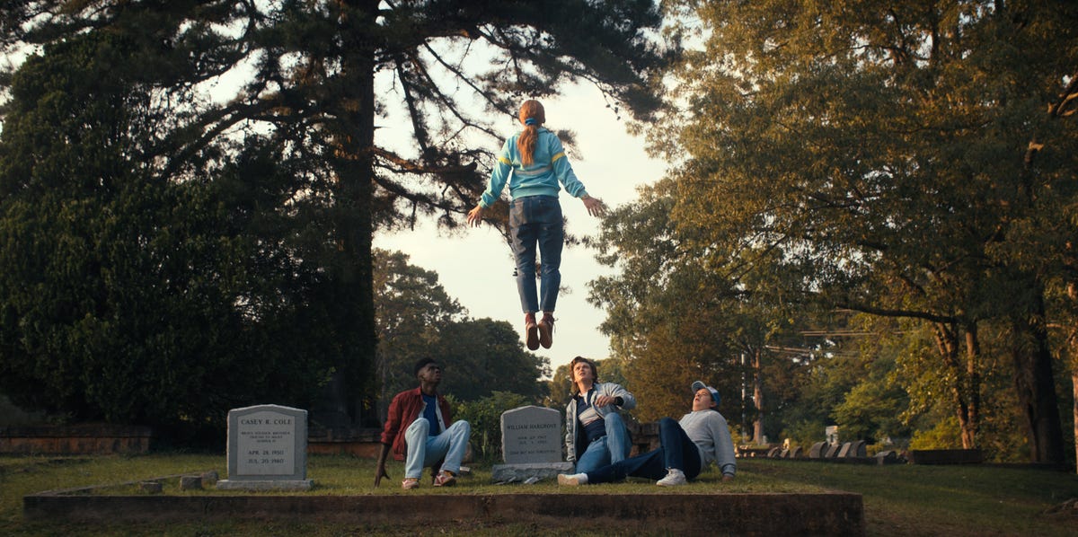 Character Max levitates above a gravestone, with other Stranger Things characters looking on in shock.
